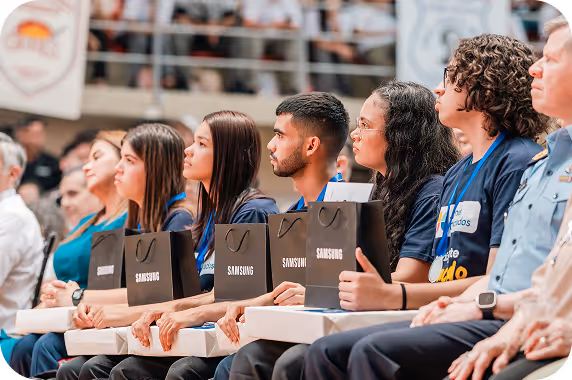 Fila de jóvenes adultos sentados, sosteniendo bolsas y cajas de regalo de Samsung en un evento bajo techo.