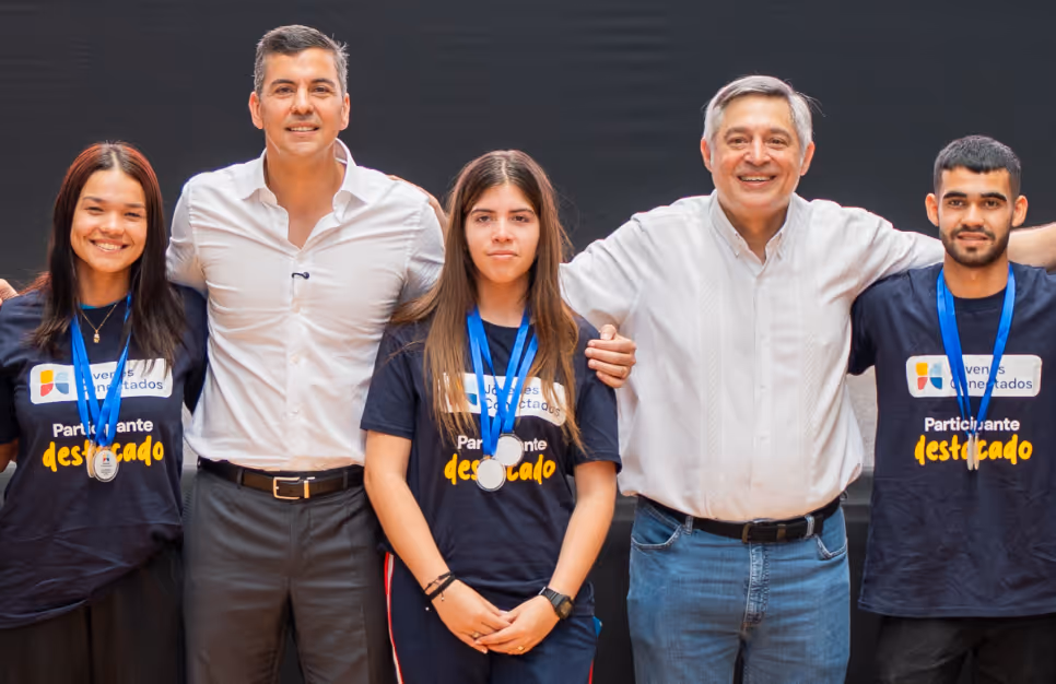 Cinco personas posando juntas contra un fondo oscuro; en el centro, hacia la izquierda, el presidente de Paraguay, Santiago Peña, junto a tres jóvenes participantes con medallas y camisetas negras, y otro hombre con camisa blanca, todos sonriendo.